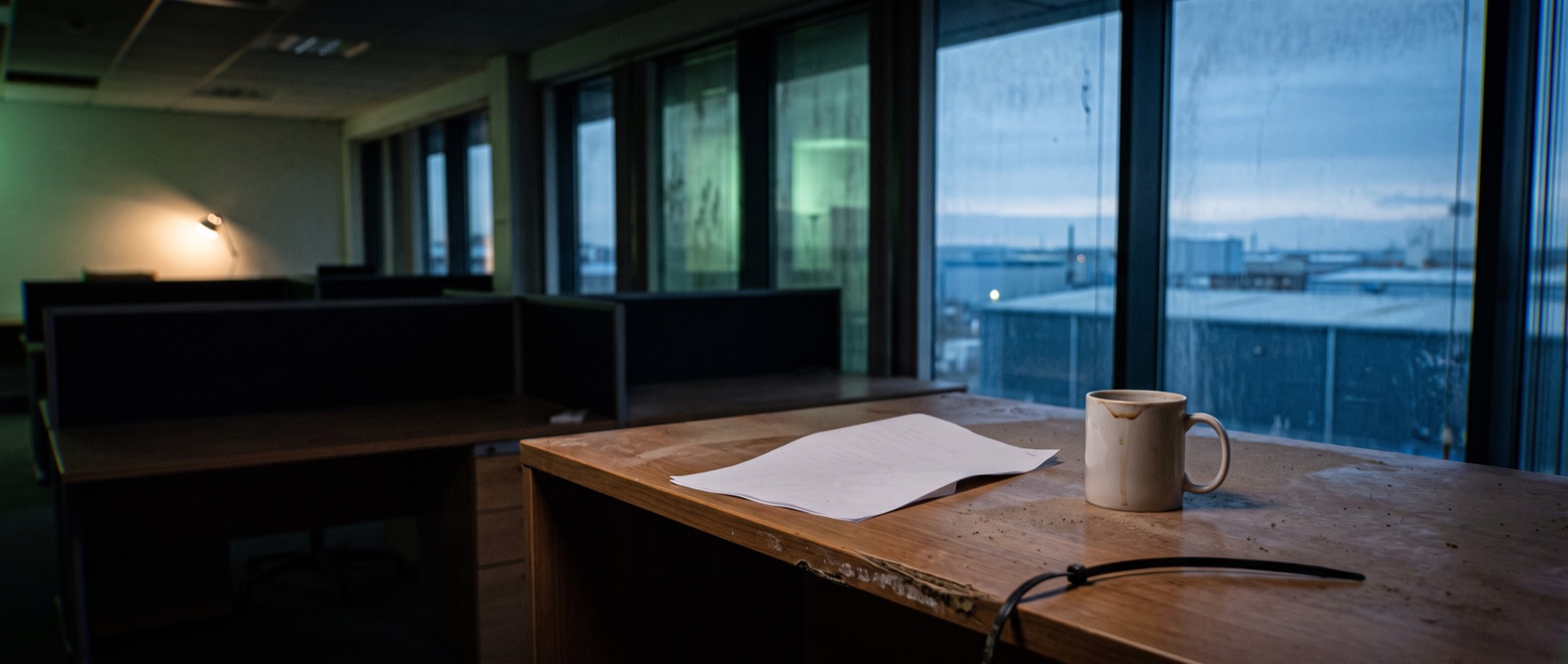 An empty open-plan office before dawn, a single sheet of paper and a cup on one desk under a lamp, sunrise over an industrial skyline beyond.