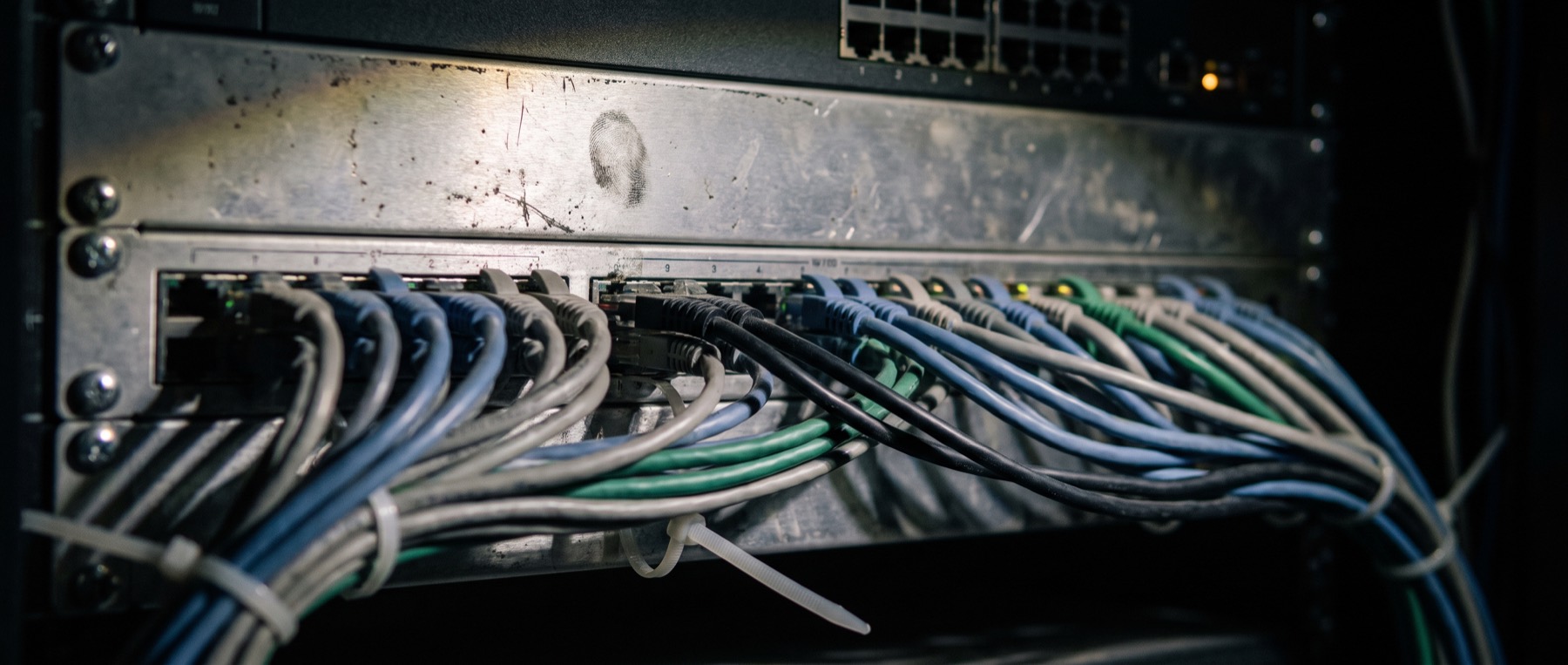 Close on a patch panel in a comms rack, rows of network leads in muted colours running into ports, dust and a fingerprint on the scratched metal face.