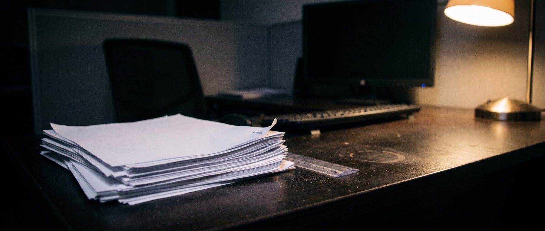 A thick stack of plain printed pages on a dark desk at night, edges slightly fanned, lit by a single lamp, a dark monitor behind.